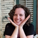 Photograph of a young white woman with brown curly hair, smiling and propping her face on her hands.